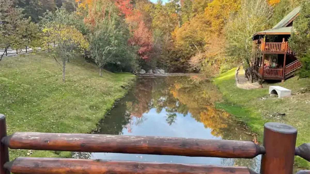 This image captures a rustic cabin overlooking calm water surrounded by vibrant, colorful foliage. A sturdy wooden railing frames the scene, inviting peaceful nature walks and moments of contemplation. The abundance of trees in their fall splendor hints at the region’s iconic scenery, and the tranquil water reflects the surrounding beauty, emphasizing the serenity of the Smoky Mountains.