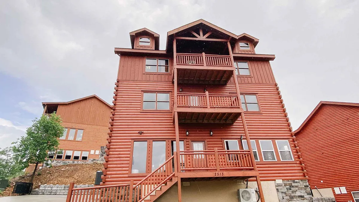 The image showcases a grand, multi-level wooden cabin nestled in the Smoky Mountains, with expansive balconies perfect for sunset viewing and spacious stairs leading to an inviting entrance. The warm glow of sunset bathes the exterior, highlighting ample outdoor spaces ideal for gatherings and relaxation.