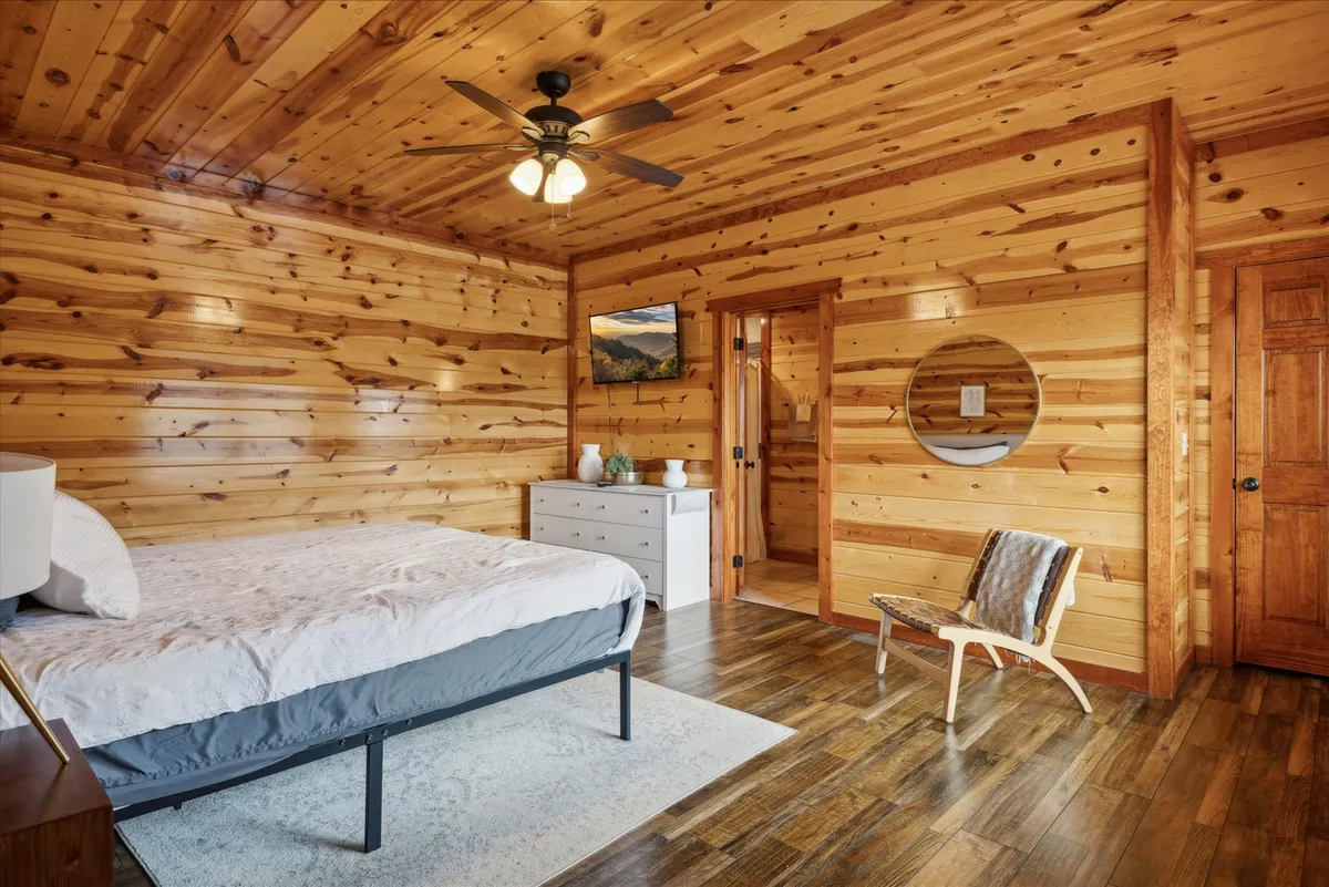 A serene bedroom with wooden wall paneling, a bed featuring simple bedding, artwork on the wall, a mirrored round frame, and a wooden chair. Natural light softly fills the space, creating a peaceful retreat ideal for relaxation.