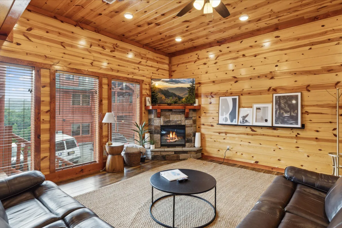 A spacious, open living room featuring a prominent stone fireplace, large windows showcasing scenic views, a coffee table, and a comfortable seating arrangement. The rustic-chic décor and abundance of natural light create a warm, inviting environment perfect for gatherings.