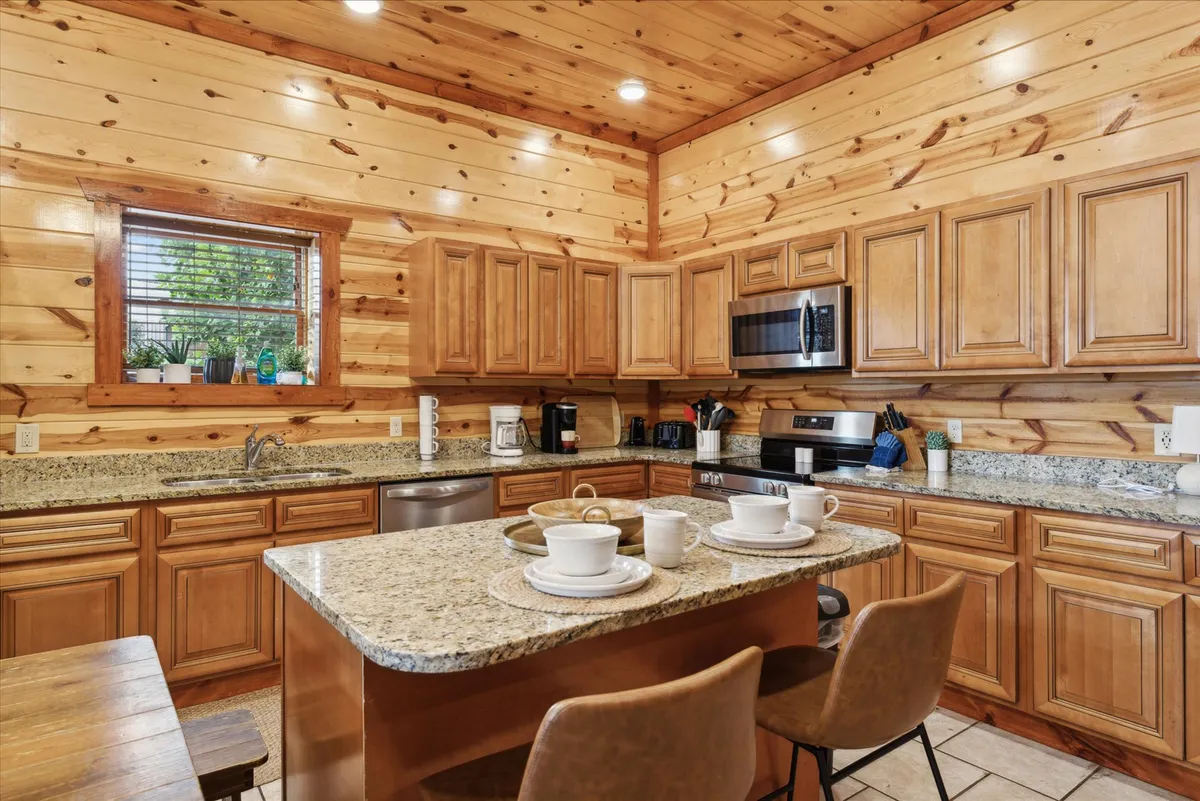 A cozy kitchen and dining area with a prominent granite countertop island, wooden cabinetry, and a welcoming dining table set beside a window with potted plants. The space exudes comfort and is ideal for gatherings, casual meals, or sharing stories while cooking.