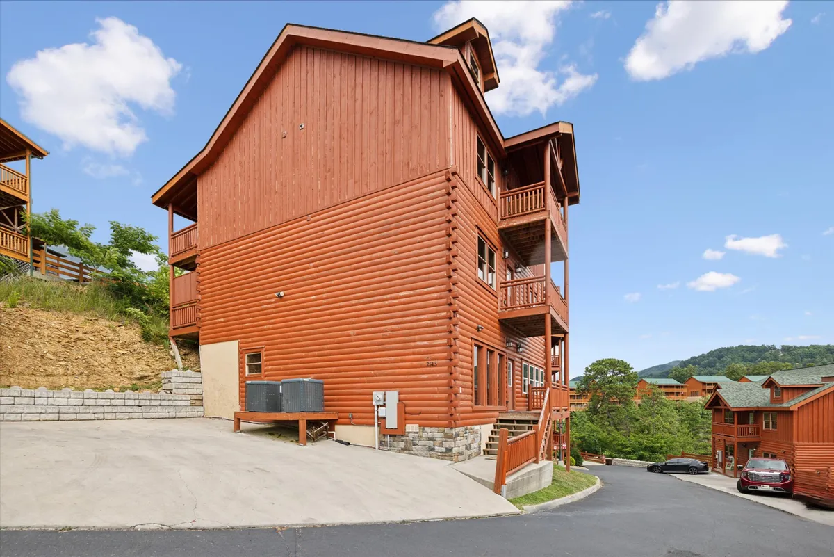 A wide view of the cabin's wooden exterior, featuring balconies overlooking lush greenery and a spacious driveway. The scene suggests opportunities for outdoor gatherings, family barbecues, and soaking in scenic views.