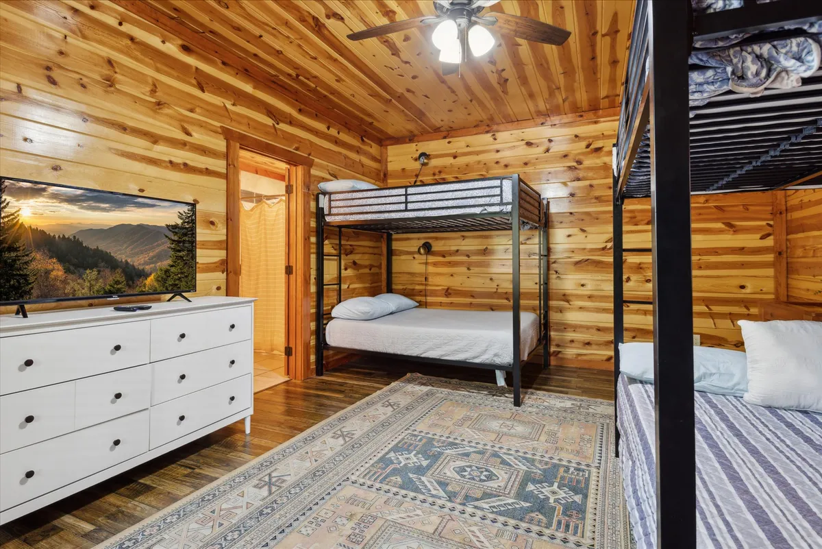 A cozy bunk room with rustic wooden walls, features sturdy bunk beds, a television, and a dresser, topped off with a playful rug. The space feels inviting and fun—perfect for kids' late-night chats and sleepovers.