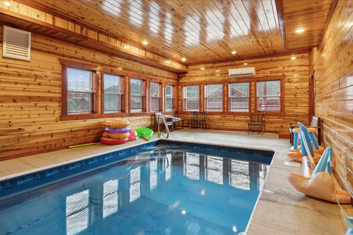 This image shows a vibrant indoor swimming pool surrounded by wooden walls. The pool is adorned with colorful pool floats, and there is a cozy seating area along the side, suggesting a space for both relaxation and fun.