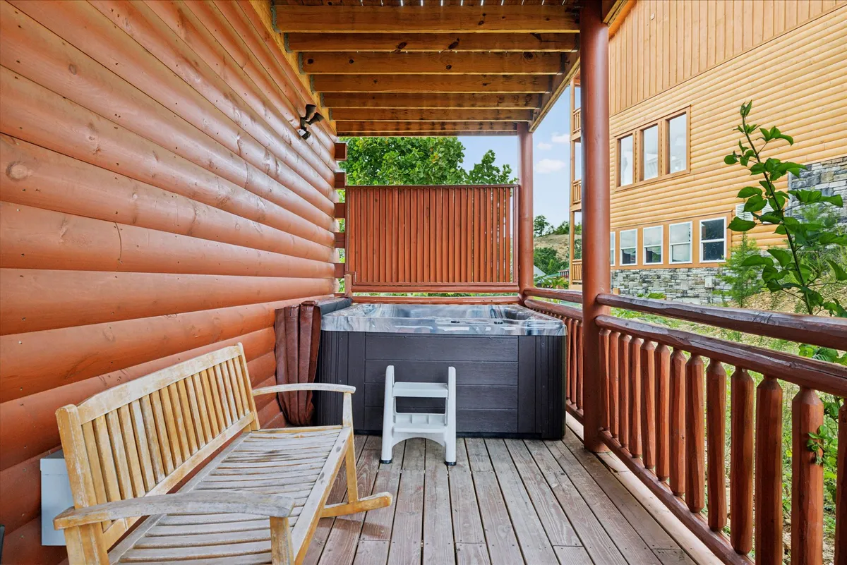 An inviting outdoor hot tub set against rustic wooden railings, accompanied by a wooden bench. The area is surrounded by natural scenery, suggesting privacy and tranquility, while subtle hints of mountain views can be seen beyond the deck edge.